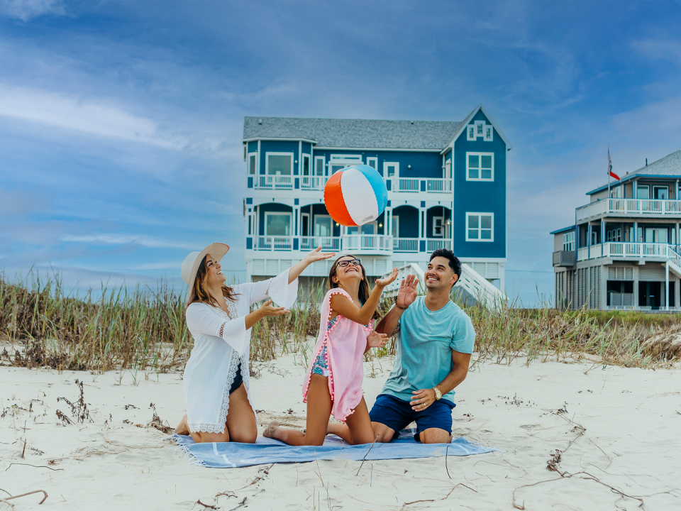 A man, woman, and young girl play with a beach ball in the sand in front of a beachfront property in Galveston, TX.