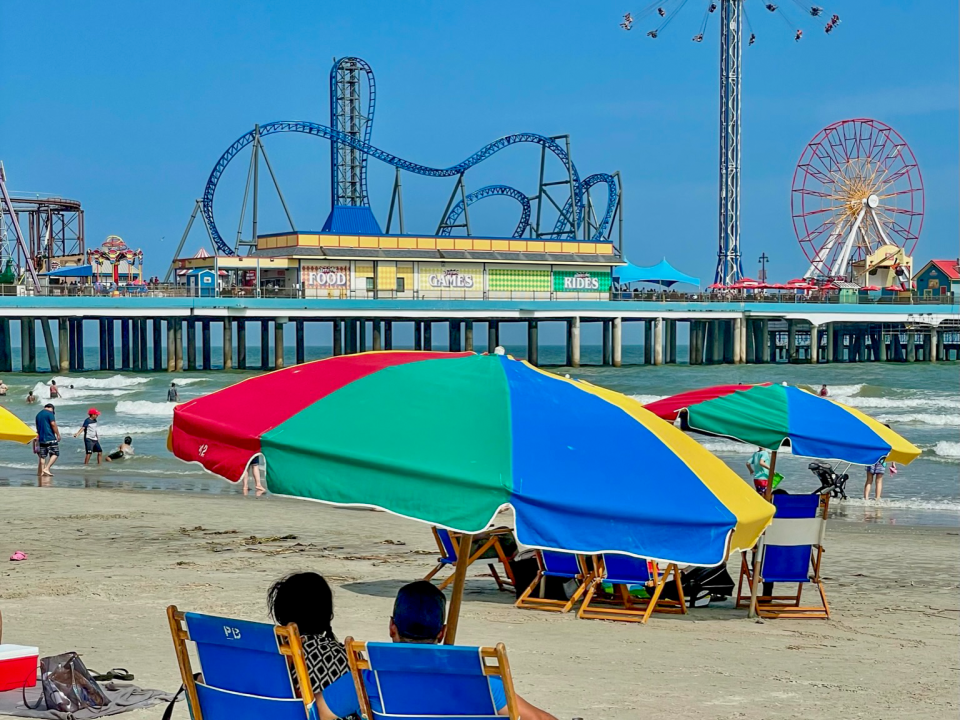 a couple sits under a colorful umbrella on the beach overlooking the ocean and Pleasure Pier in Galveston, Texas