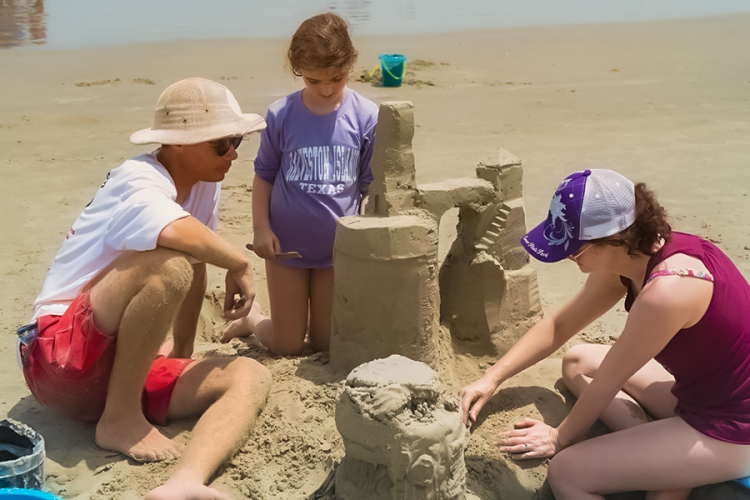 Three beachgoers build a sandcastle on the beach in Galveston Island, TX.