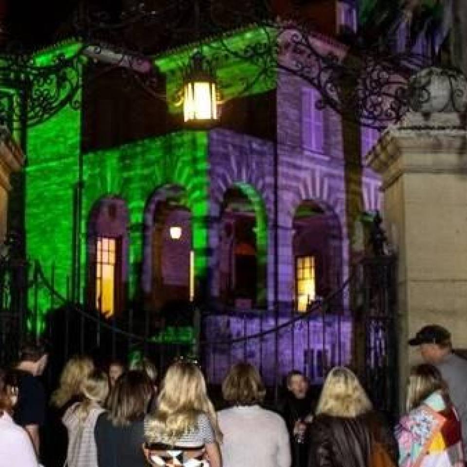 People stand in front of a large iron gate admiring a building illuminated with green and purple lights on a ghost tour