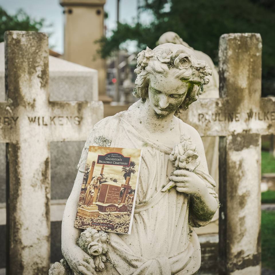 A book about Galveston cemeteries leans against a statue of a female figure during a cemetery tour in Galveston
