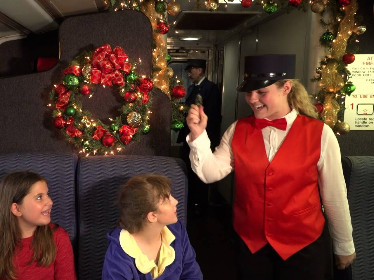 two children smile up at a train conductor while sitting under a wreath decorated with red and green ornaments on a train