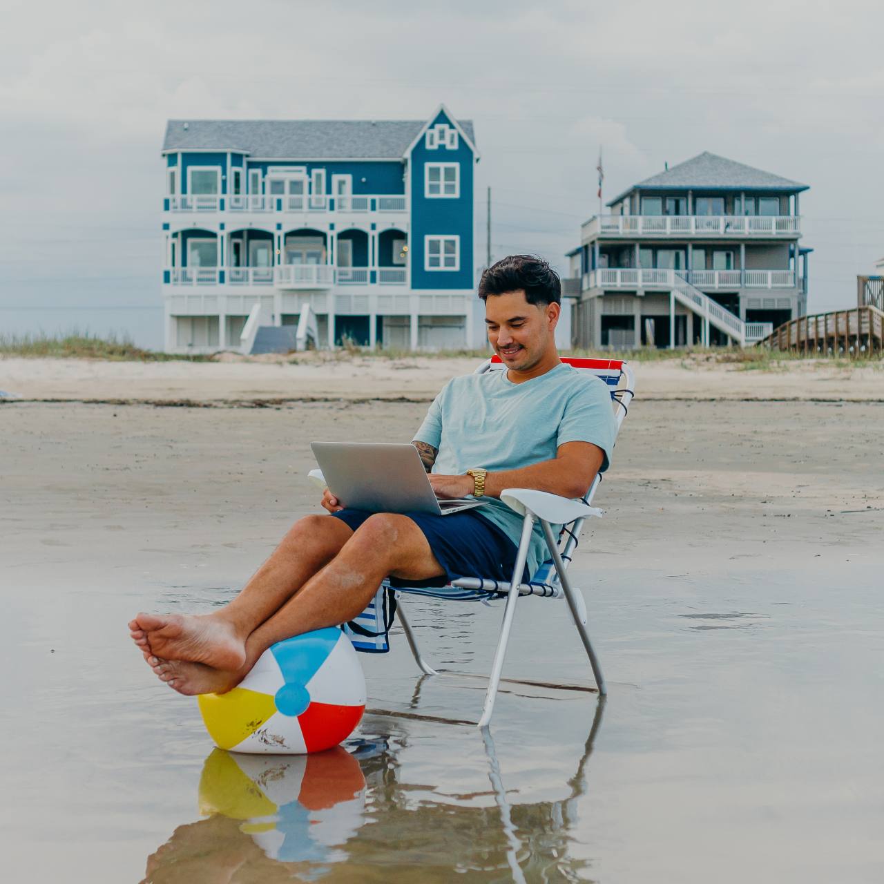 a man sitting on the beach in a beach chair with his legs on a beach ball while working on a laptop in Galveston