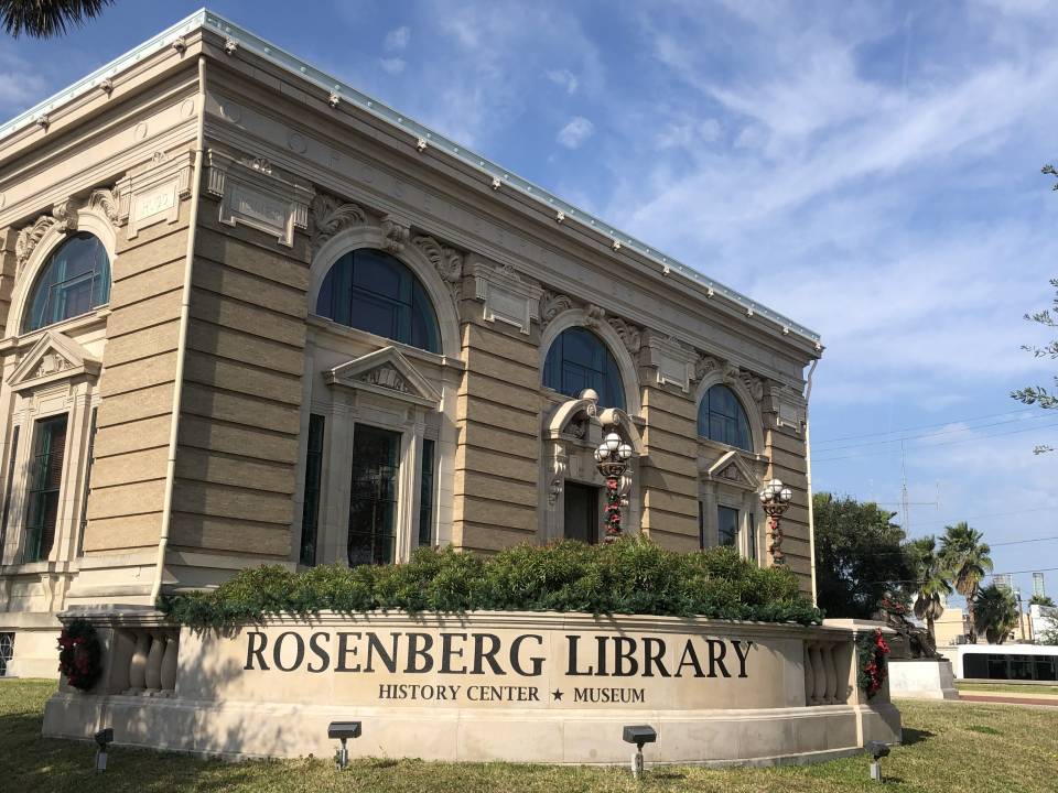 The exterior of Rosenberg Library surrounded by bushes and standing globe lights.
